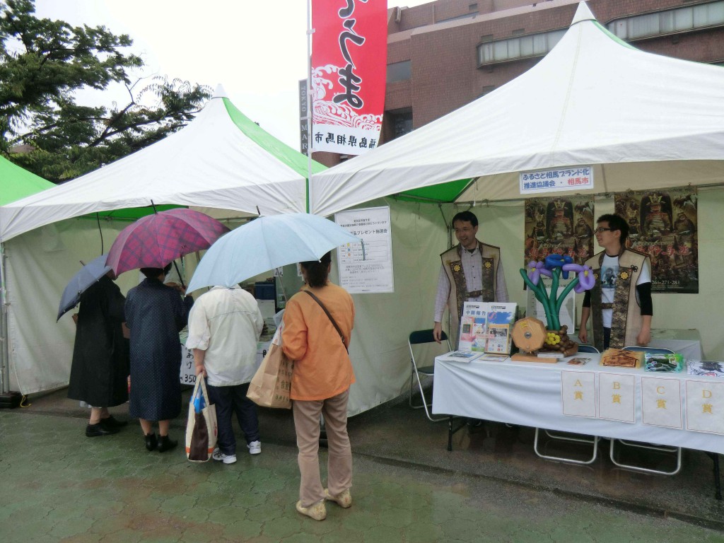 ・傘を差し、海鮮フーズのブースに来場してくださったお客様たち。お隣がふるさと相馬ブランド化推進協議会のブースです。